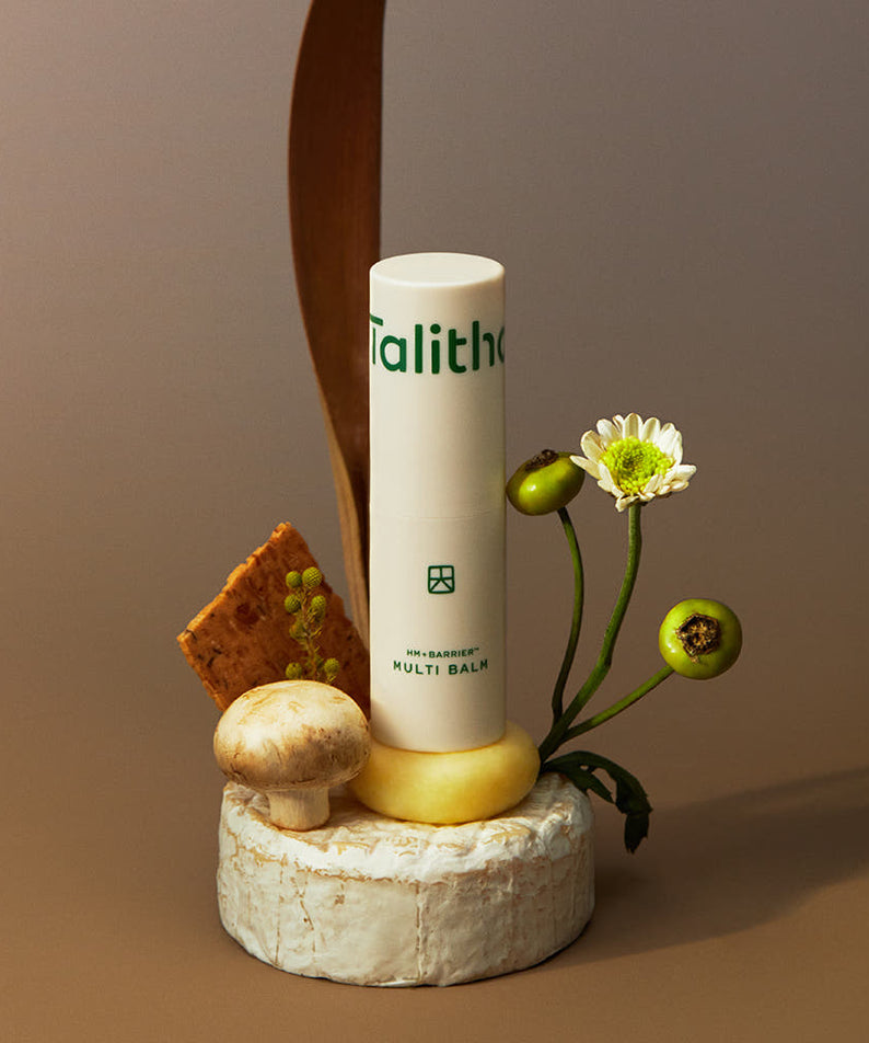 TALITHA KOUM HM+ Barrier Multi Balm 9g de Talitha, sur un socle avec ingrédients naturels et fleurs, fond beige.