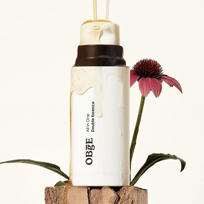 OBGE All-in-one double essence 100ml, bouteille cosmétique OBGE sur pierre avec fleur rouge, fond clair (photo produit)