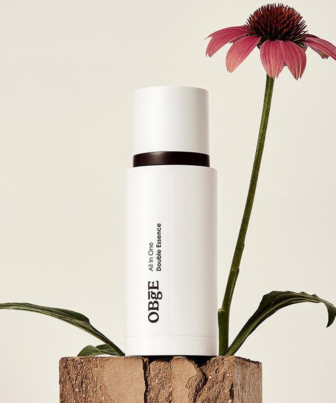 OBGE All-in-one double essence 100ml : flacon blanc OBGE sur socle bois, fleur violette en décor naturel, fond clair.