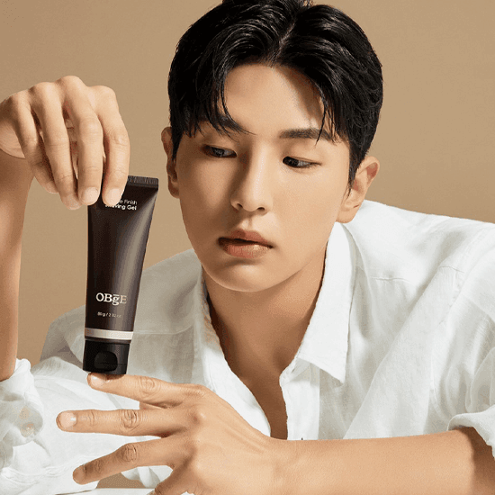 Décrivez l'image : OBGE Gentle Finish Shaving Gel 80g, flacon tenu par un homme en préparation de rasage, fond beige.