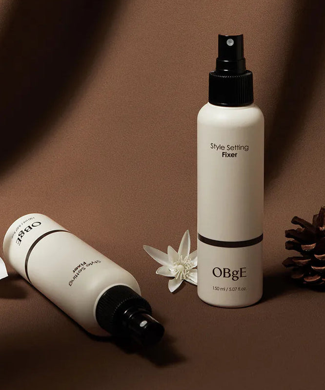 OBGE Style Setting Fixer 150ml, brume coiffante OBGE sur fond beige, avec fleurs et accessoires décoratifs.
