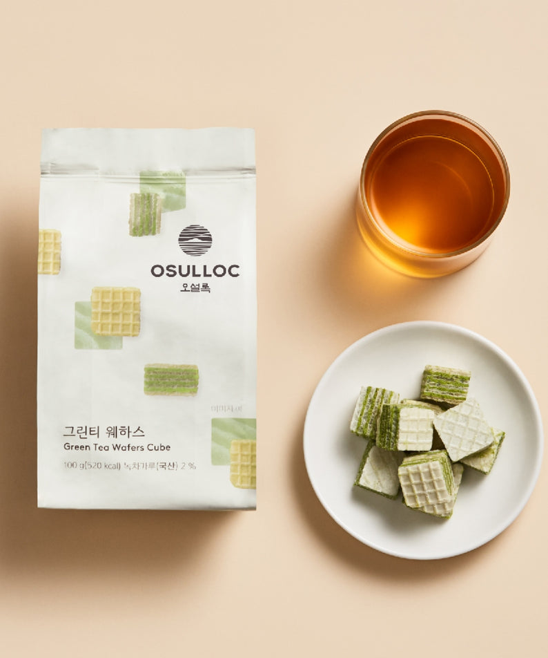 Décris l'image en français : OSULLOC Green Tea Wafers 100g, sachet et gaufres vertes avec tasse de thé sur fond clair.