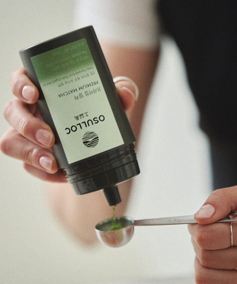 OSULLOC Premium Matcha 40g : main versant du matcha, pot vert OSULLOC sur table, contexte cuisine/boisson.