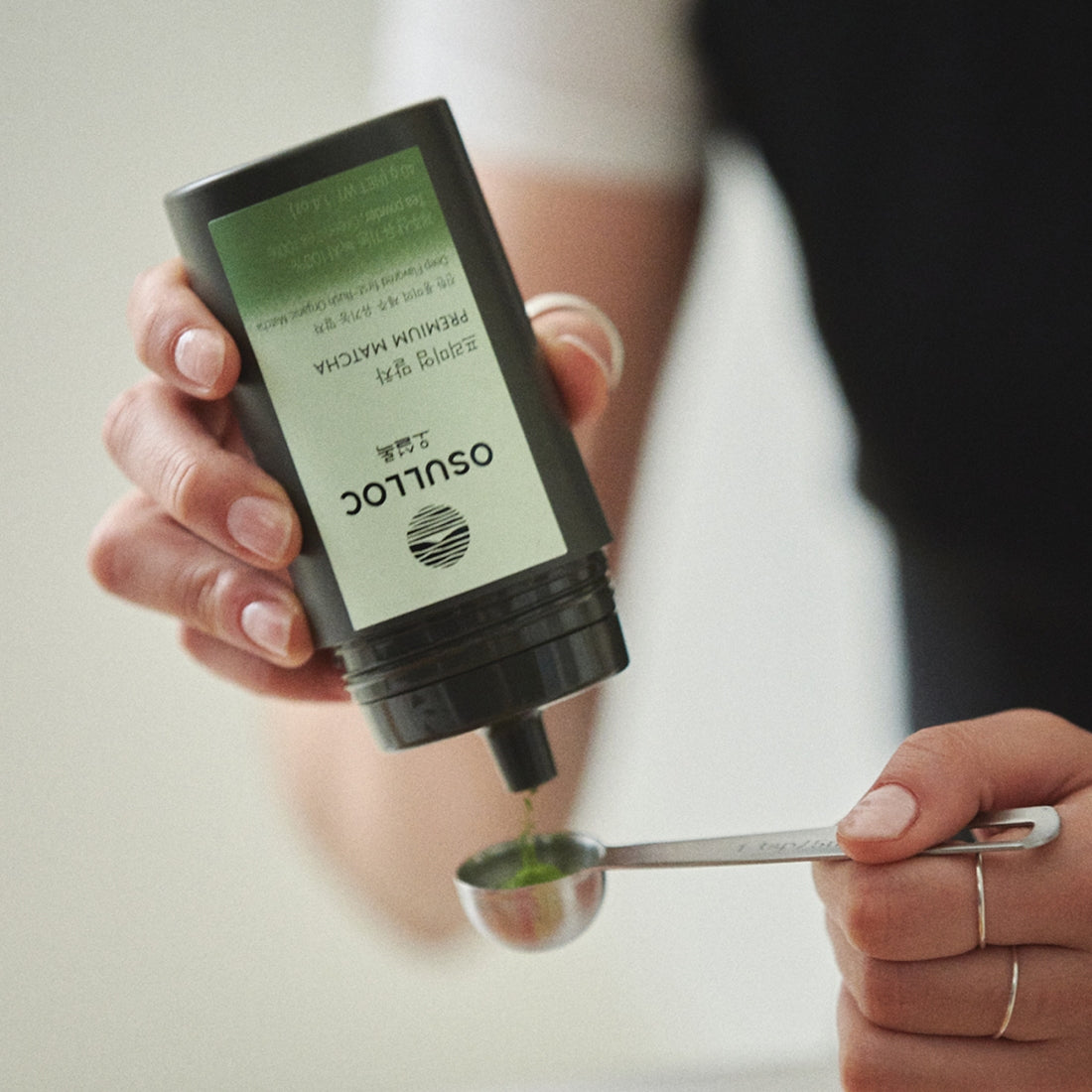 OSULLOC Premium Matcha 40g : main versant du matcha, pot vert OSULLOC sur table, contexte cuisine/boisson.