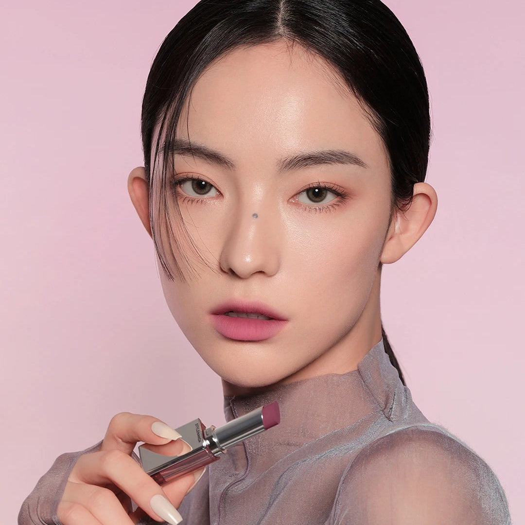 Modèle maquillée appliquant la 3CE Blur Matte Lipstick (3CE) sur fond rose, rendu lèvres mates.