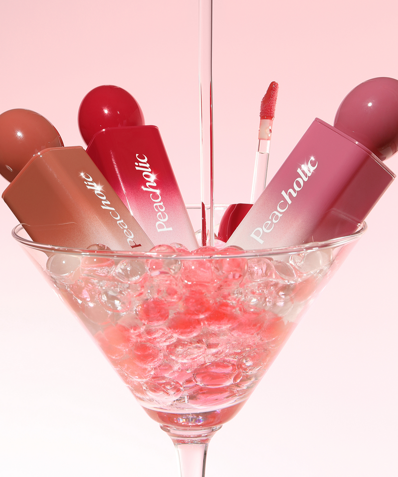 PEACHOLIC Water Glow Lip Tint de Peacholic, maquillage en gouttelettes roses, tube et gloss en flûte sur fond rose.