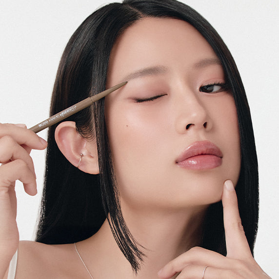 Décris l'image en français : PERIPERA Speedy Eyebrow Auto Pencil 25AD, main applique un crayon sur les sourcils, femme maquillée.