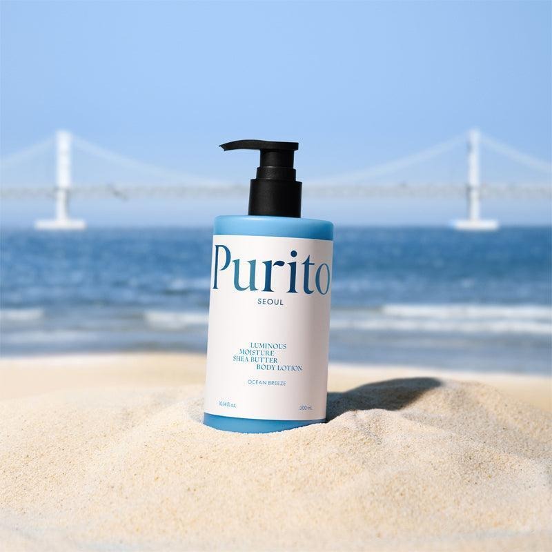 PURITO Luminous Moisture Shea Butter Body Lotion 300ml, flacon sur une plage en bord de mer, produit Purito en contexte visuel.