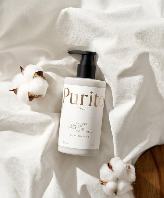 PURITO Luminous Moisture Shea Butter Body Lotion 300ml, flacon Purito sur tissu blanc avec coton, ambiance spa.
