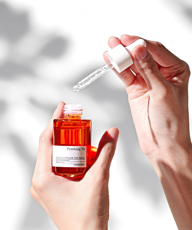 Flacon de PYUNKANG YUL Brightening Blemish Care Serum 30ml tenu dans une main, avec une pipette appliquant le sérum en lumière naturelle.