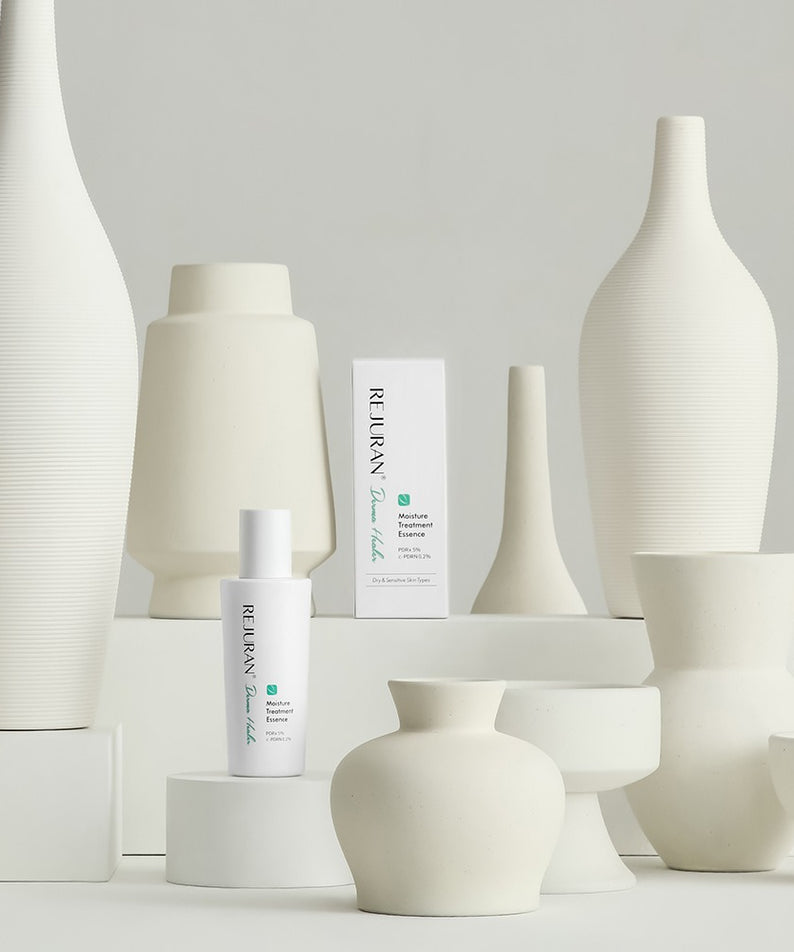 Décris l'image en français : REJURAN Derma Healer Moisture Treatment Essence 70g, flacons blancs sur décor minimal, marque REJURAN.