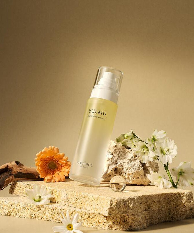 Flacon de RETURNITY Yulmu Calming Toner Mist 100ml entouré de fleurs, posée sur une pierre naturelle beige.