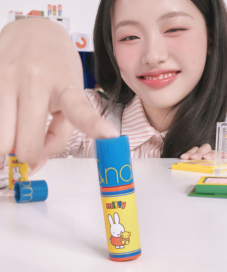 ROMAND  Glasting Color Gloss MINI de la marque ROMAND, tube maquillage sur bureau, maquillage studio, près d’une femme souriante.