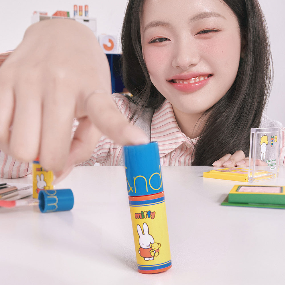 ROMAND  Glasting Color Gloss MINI de la marque ROMAND, tube maquillage sur bureau, maquillage studio, près d’une femme souriante.
