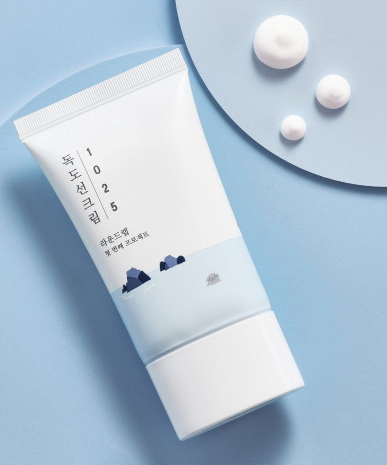 ROUND LAB 1025 Dokdo Sun Cream 50ml sur fond bleu ciel minimaliste avec texture de crème solaire, packaging élégant.