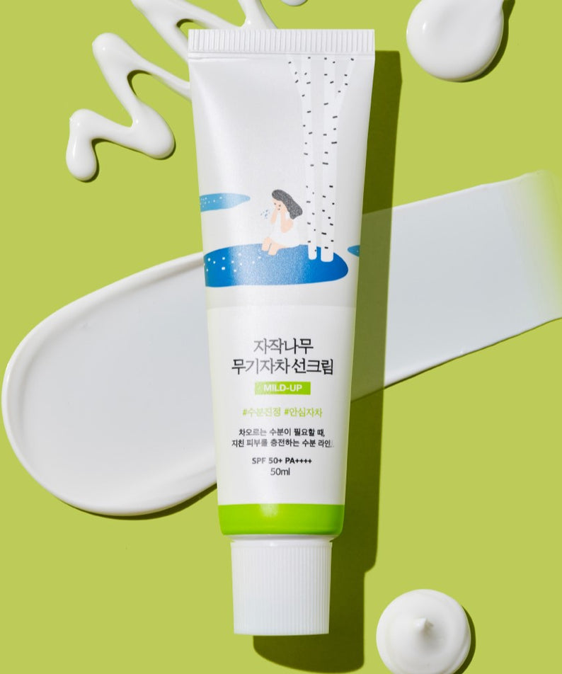 Crème solaire ROUND LAB Birch Juice Moisturizing Mild-Up Sunscreen 50ml sur fond vert avec textures de crème.