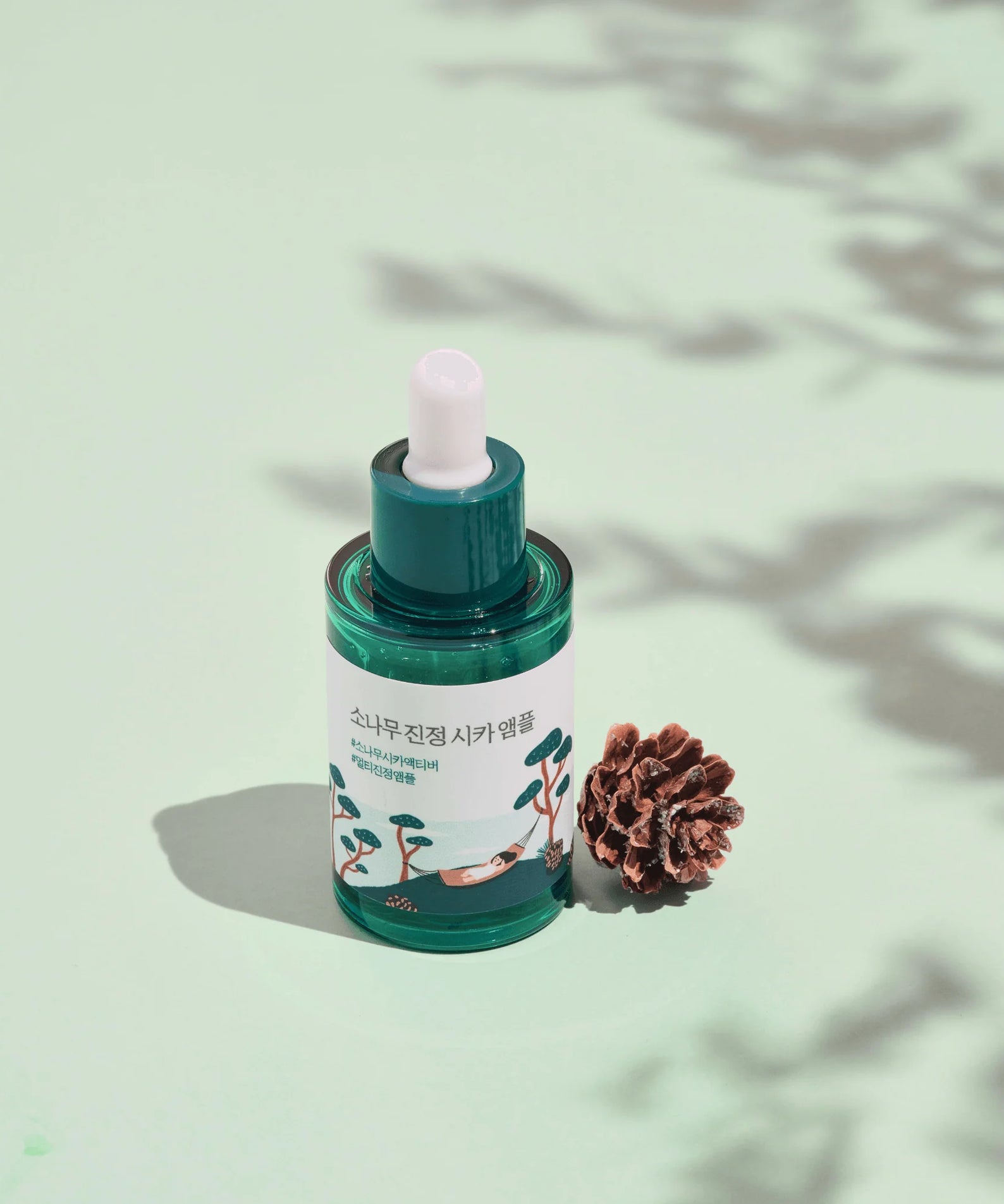 ROUND LAB Pine Calming Cica Ampoule 30ml posée à côté d'une pomme de pin sur fond vert, sérum apaisant pour peau sensible.