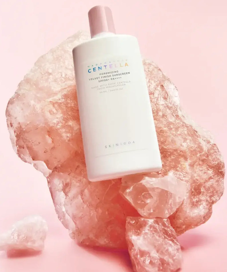 SKIN1004 Madagascar Centella Poremizing Velvet Finish Sunscreen 50ml, flacon crème, sur fond rose avec pierre minérale.
