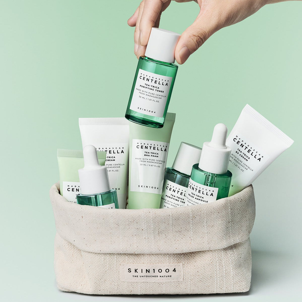 Décris l'image en français: SKIN1004 Madagascar Centella Teatrica Travel Kit de SKIN1004, trousses dans une pochette blanche, fond vert.