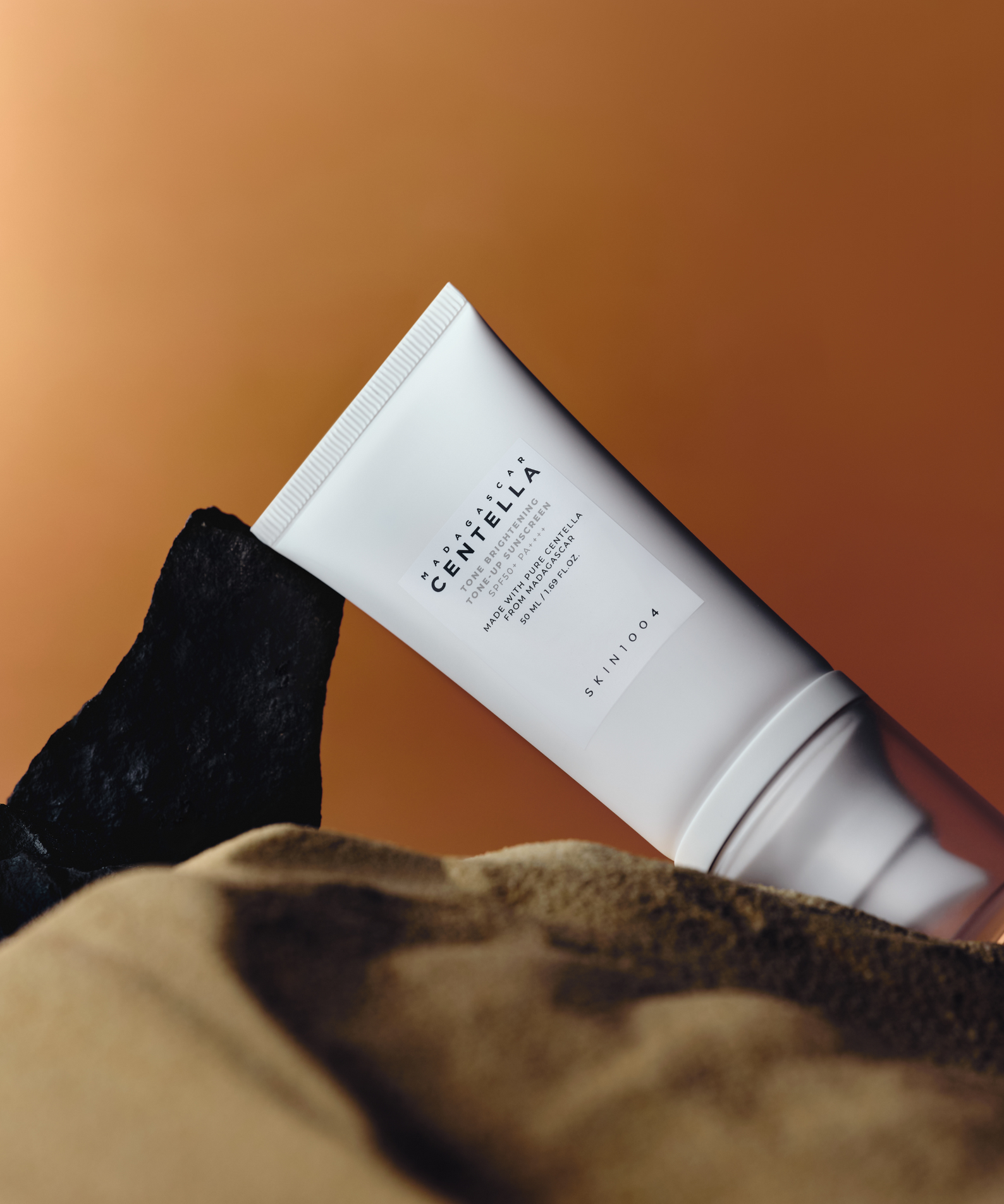 Crème solaire SKIN1004 Madagascar Centella Tone Brightening Tone-Up Sunscreen 50ml sur fond de couleur sable et noir.