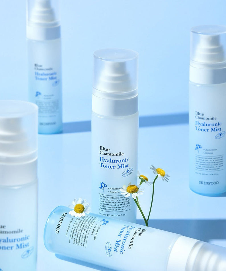 Décris l'image en français: SKINFOOD Blue Chamomile Hyaluronic Toner Mist 100ml, flacons blancs sur fond bleu avec fleurs de camomille.