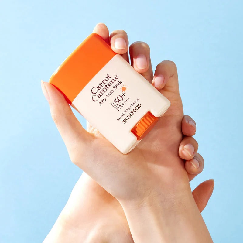 SKINFOOD Carrot Airy Soft Sun Stick 18.5g, marque SKINFOOD, tube crème solaire tenue sur fond bleu clair.