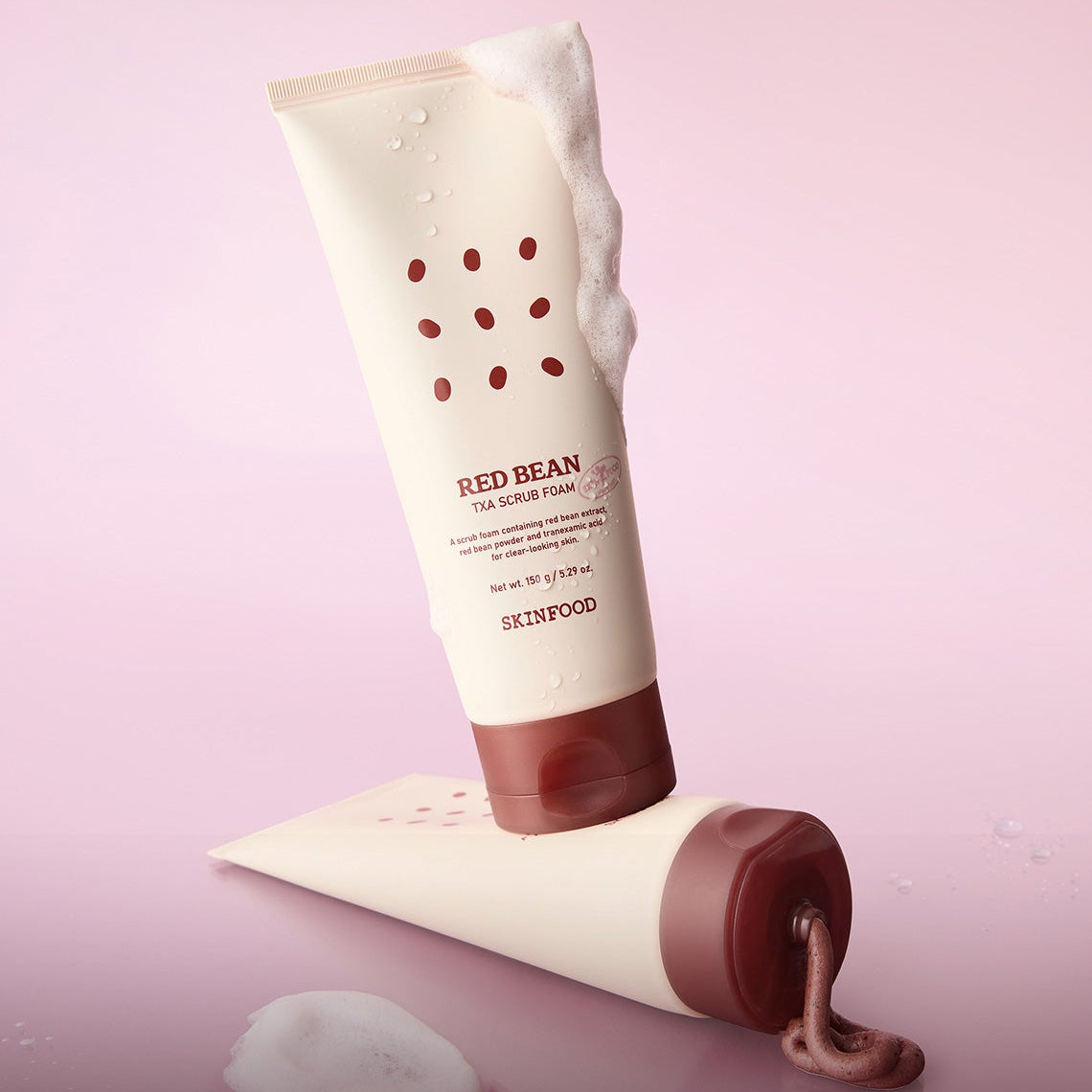 Décris l'image en français : SKINFOOD Red Bean Tranexamic Acid Scrub Foam 150g, tube et mousse sur fond rose.