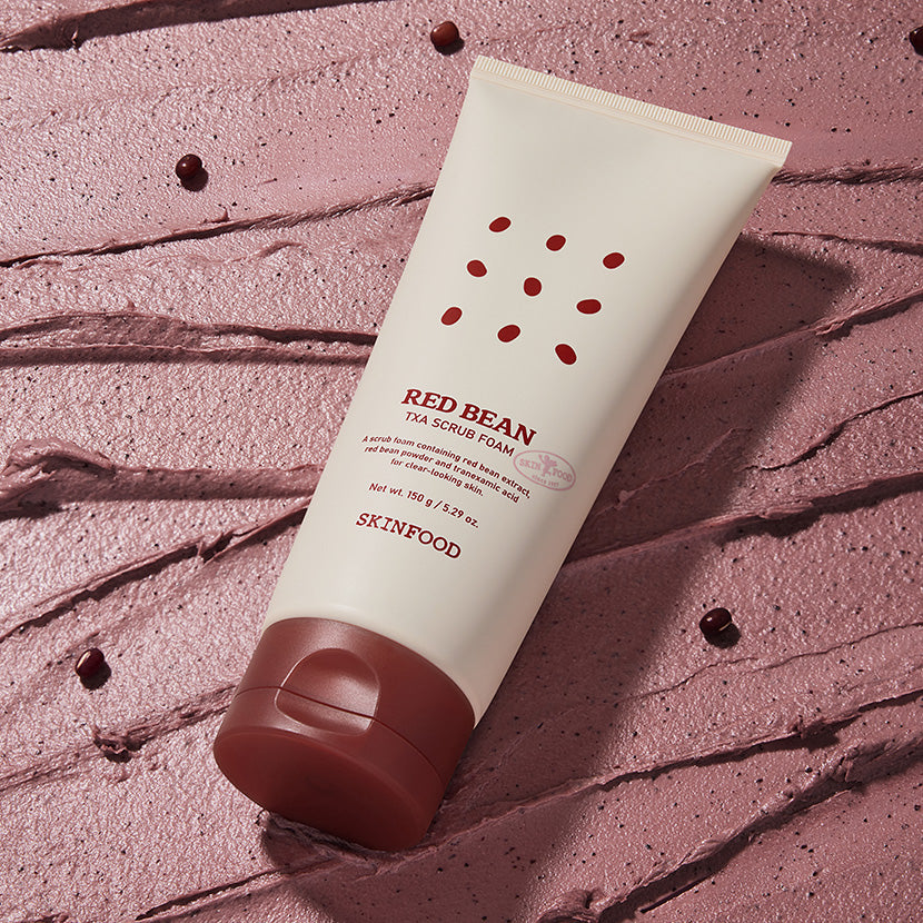 SKINFOOD Red Bean Tranexamic Acid Scrub Foam 150g en tube sur fond rose texturé, marque SKINFOOD, contexte visuel beauté.