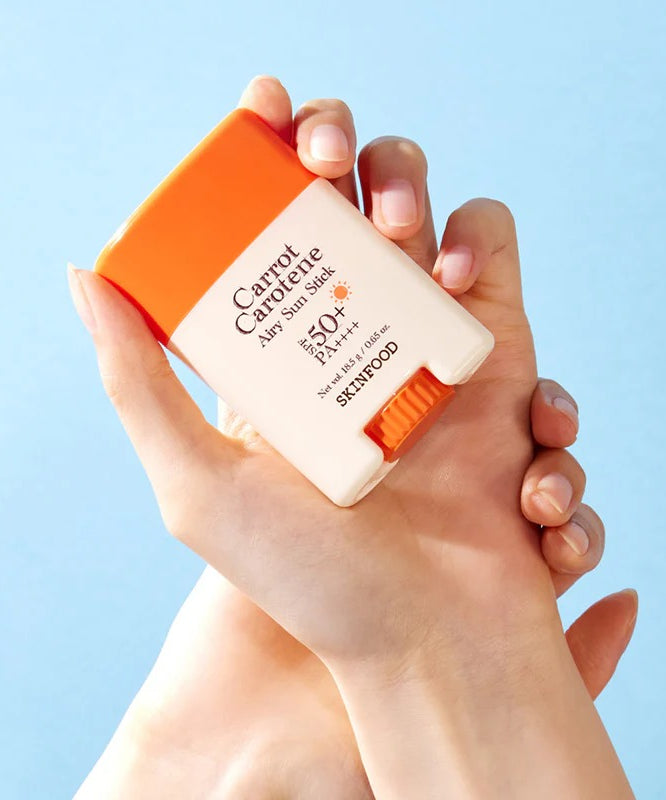 SKINFOOD Carrot Airy Soft Sun Stick 18.5g tenu dans les mains, produit solaire SPF50+ efficace et léger, fond bleu.