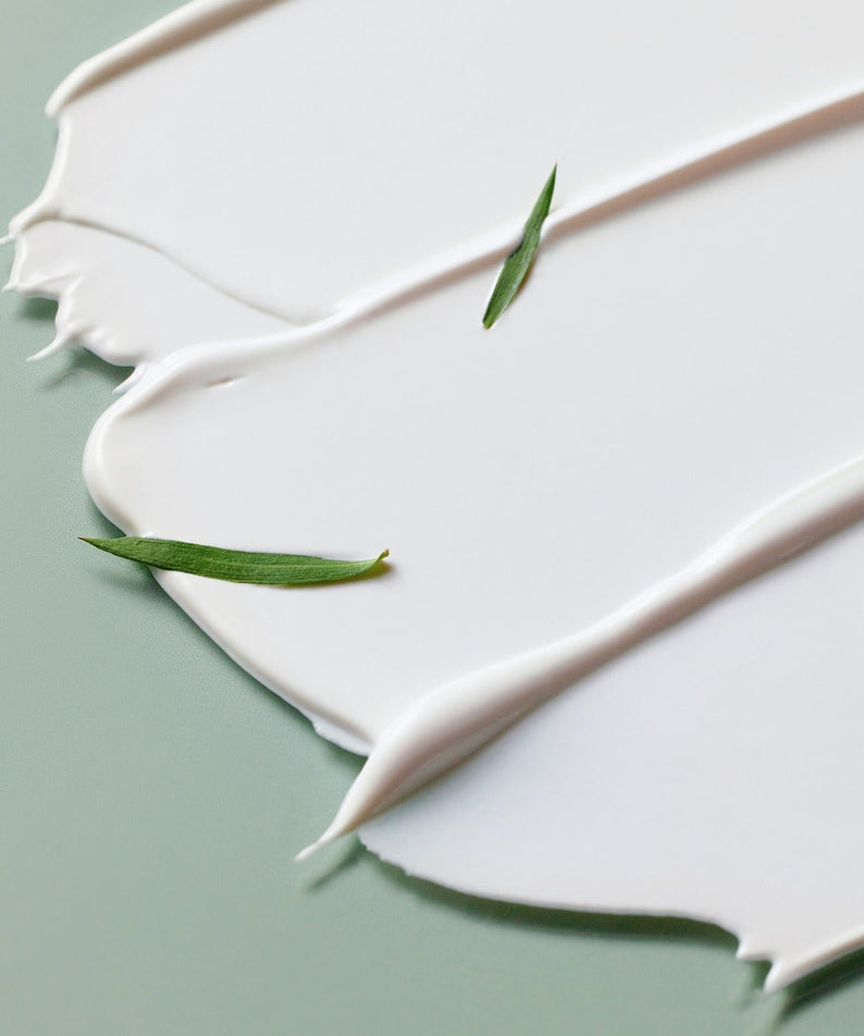 Texture de la SOME BY MI Truecica Weaponless Calming Tone Up Sun Cream 50ml sur une surface avec des feuilles de thé.