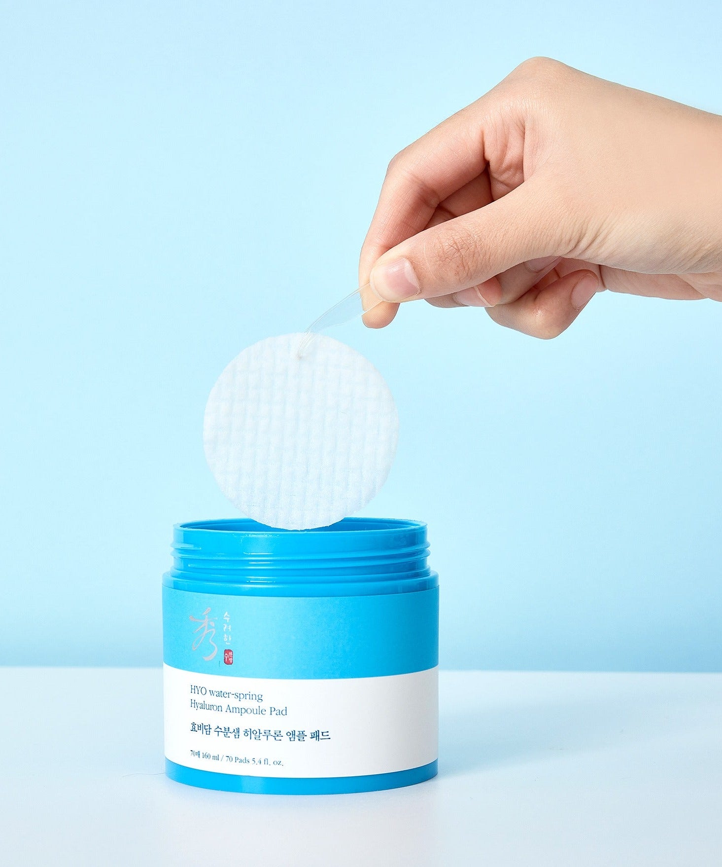 Main tenant un tampon de SOORYEHAN Moiture Spring Hyaluronic Ampoule Pads 70 Sheets au-dessus du pot bleu ouvert.