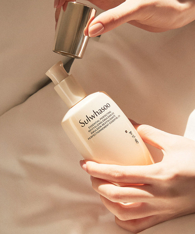 Décrivez l'image en français : SULWHASOO Essential Revitalizing Emulsion Ex 125ml, mains appliquant l'émulsion sur peau, fond beige.