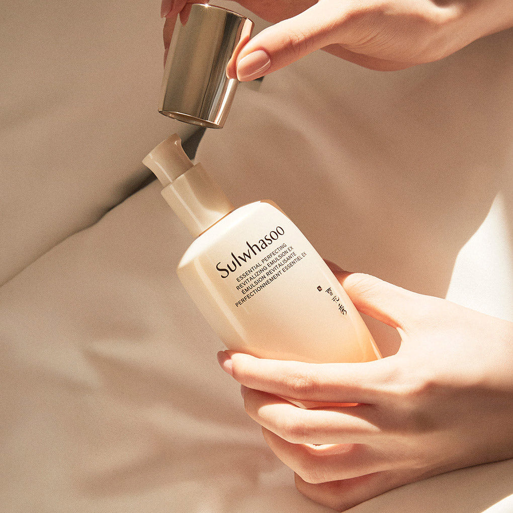 Décrivez l'image en français : SULWHASOO Essential Revitalizing Emulsion Ex 125ml, mains appliquant l'émulsion sur peau, fond beige.