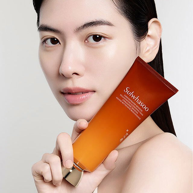 Portrait avec SULWHASOO Ginseng Rejuvenating Cleansing Foam 150g de Sulwhasoo, tube orange en main, fond clair.