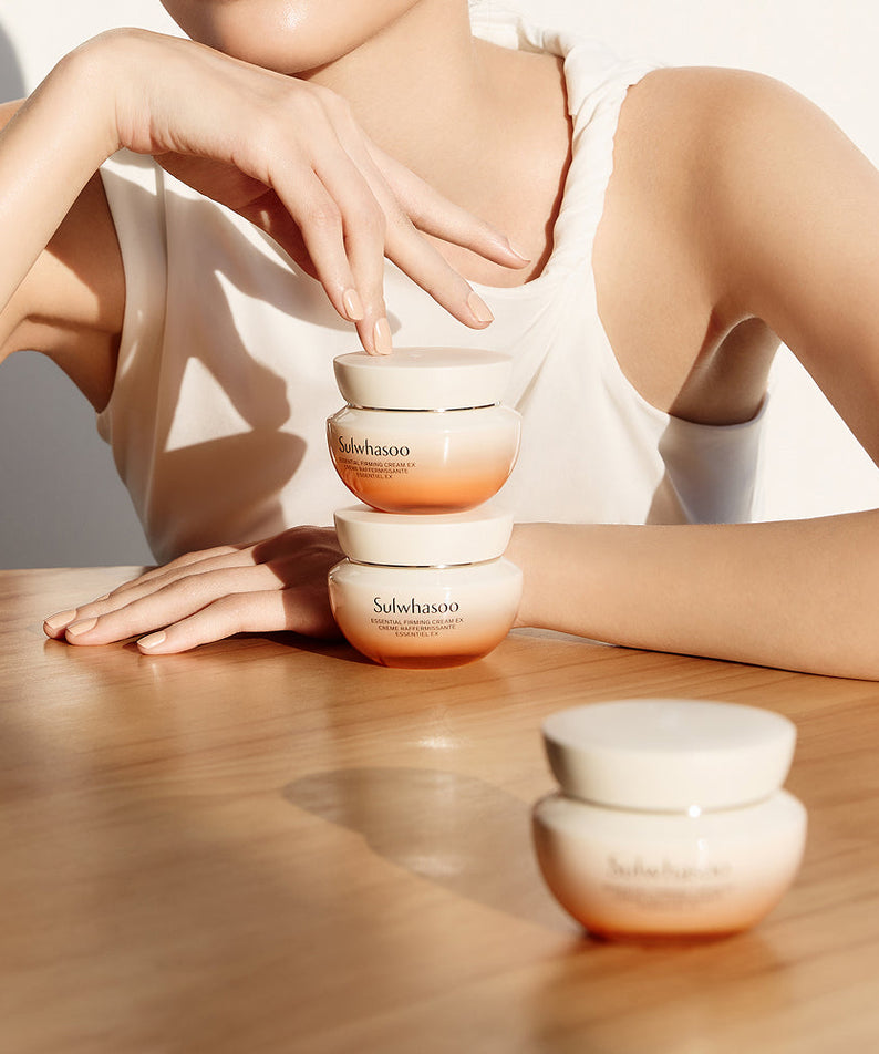 Femme posant avec SULWHASOO Essential Comfort Firming Cream 75ml, trois pots de crème sur une table en bois.