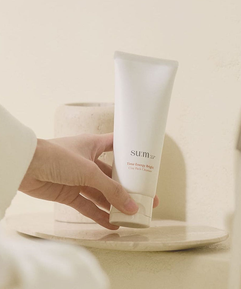 Femme tenant un tube de SU:M37 Time Energy Bright Clay Pack Cleanser 150ml sur un évier dans une salle de bain chic.