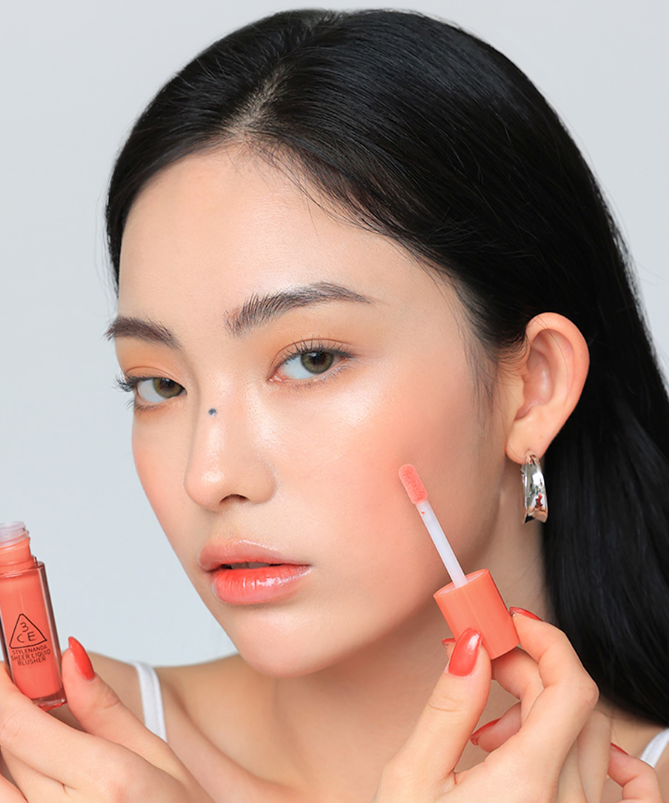 Décris l'image en français : 3CE Sheer Liquid Blusher sur une femme, application de blush liquide corail par la marque 3CE.