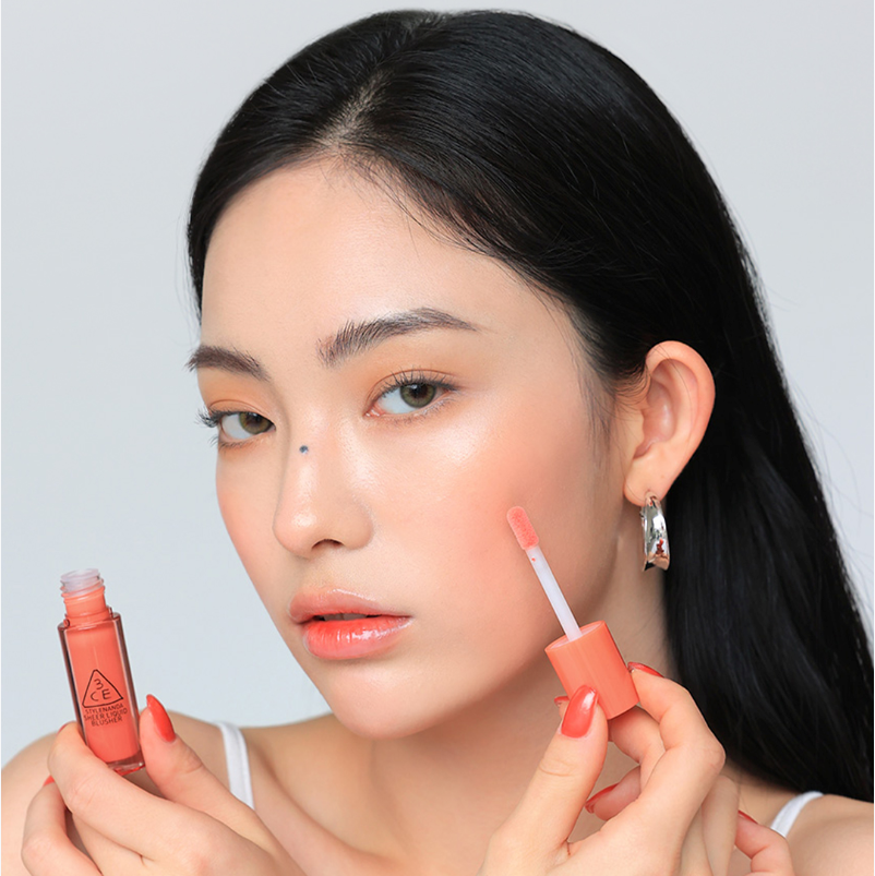 Décris l'image en français : 3CE Sheer Liquid Blusher sur une femme, application de blush liquide corail par la marque 3CE.