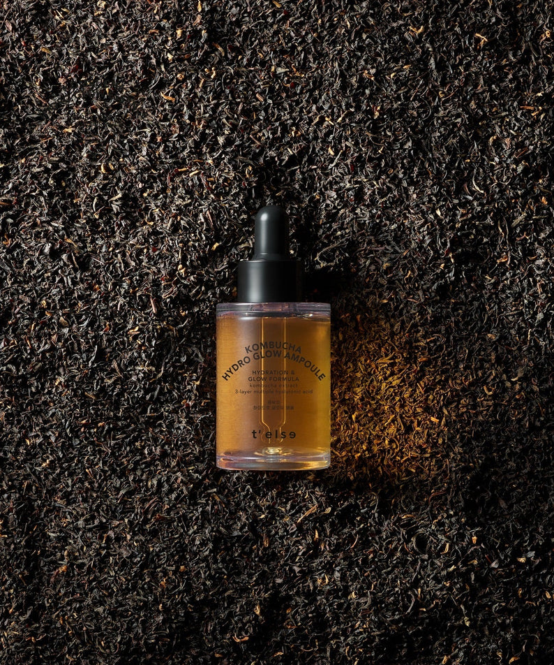 Photo de l'ampoule T'ELSE Kombucha Hydro Glow 30ml posée sur un fond de feuilles de thé noir. Cosmétique hydratante pour un teint éclatant.