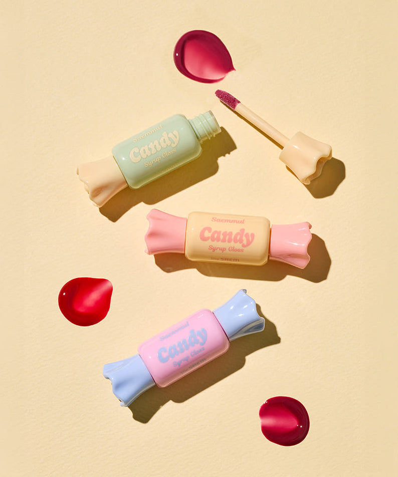 Produits de beauté THE SAEM Saemmul Candy Syrup Gloss exposés avec des gélules colorées sur un fond beige.