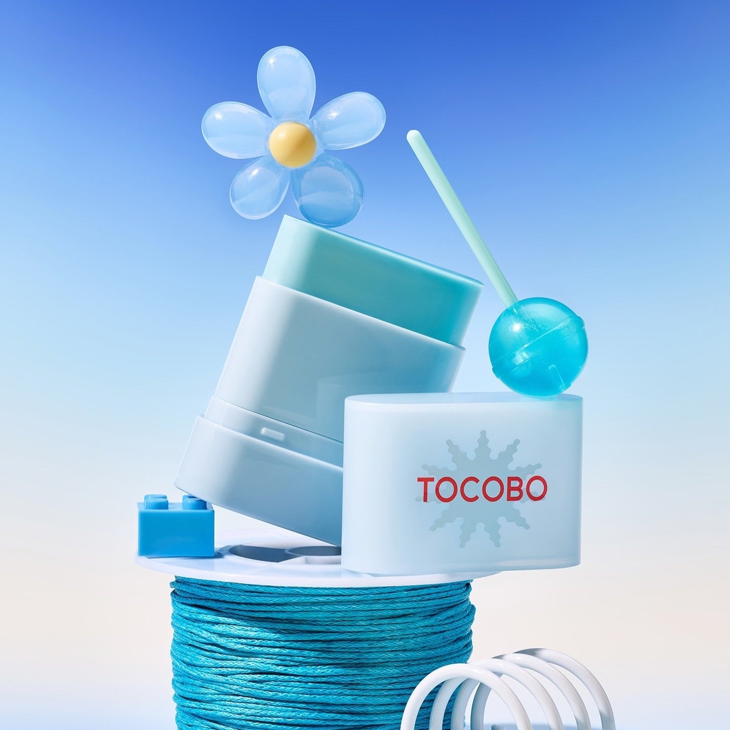 TOCOBO Mini Sun Stick Trio de TOCOBO, en pack sur fond bleu ciel avec accessoires visuels, fleur et soleil stylisés.