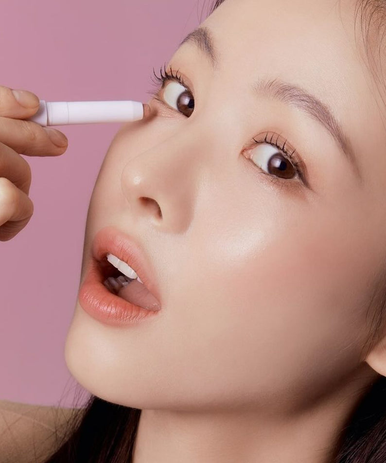 Femme appliquant TONY MOLY The Shocking Color Fixing Stick Shadow sur paupière, maquillage subtil et lumineux sur fond rose.