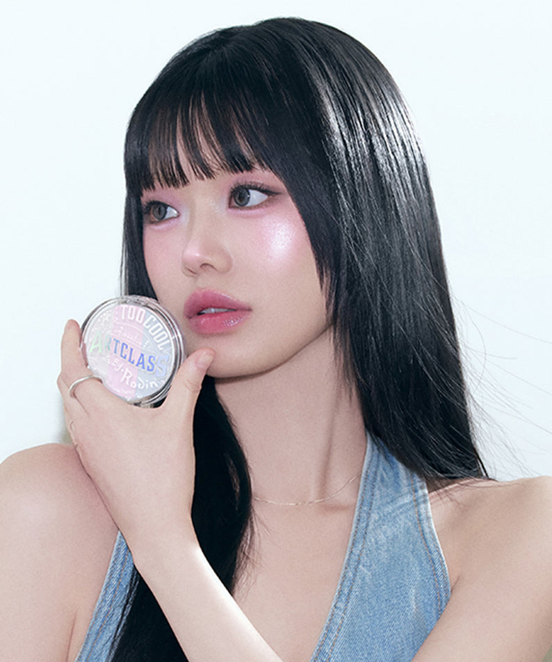 Femme tenant le TOO COOL FOR SCHOOL Art Class By Rodin Highlighter sur sa joue, lumière douce, cheveux noirs brillants, maquillage éclatant.