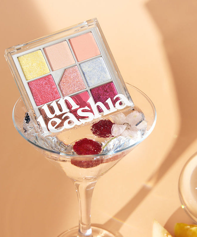 UNLEASHIA Glitterpedia Eye Palette, palette fards pailletés en coffret transparent sur fond beige, gros plan en lumière chaude.