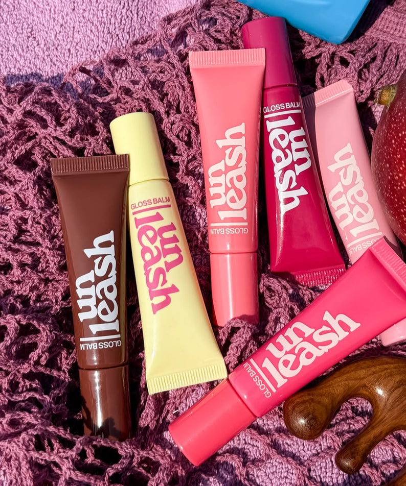 Collection de baumes UNLEASHIA Sunset Dazzle Gloss Balm sur texture rose.
