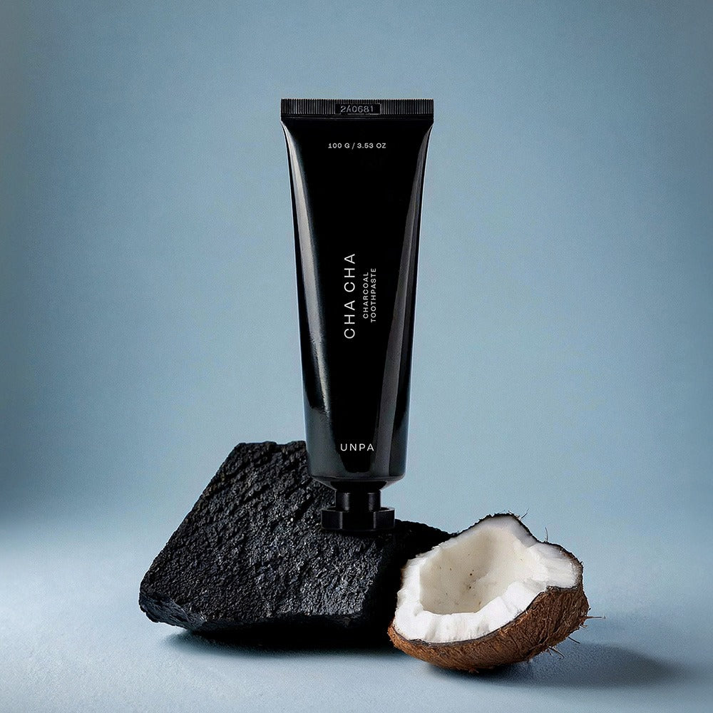 UNPA Cha Cha Charcoal Original Toothpaste 100g de UNPA : tube noir sur charbon et noix de coco, fond bleu.