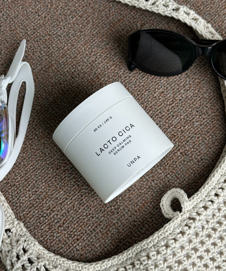 UNPA Lacto Cica Deep Calming Serum Pad 85 Sheets UNPA, pot blanc sur tissu beige, lunettes et accessoires en arrière-plan