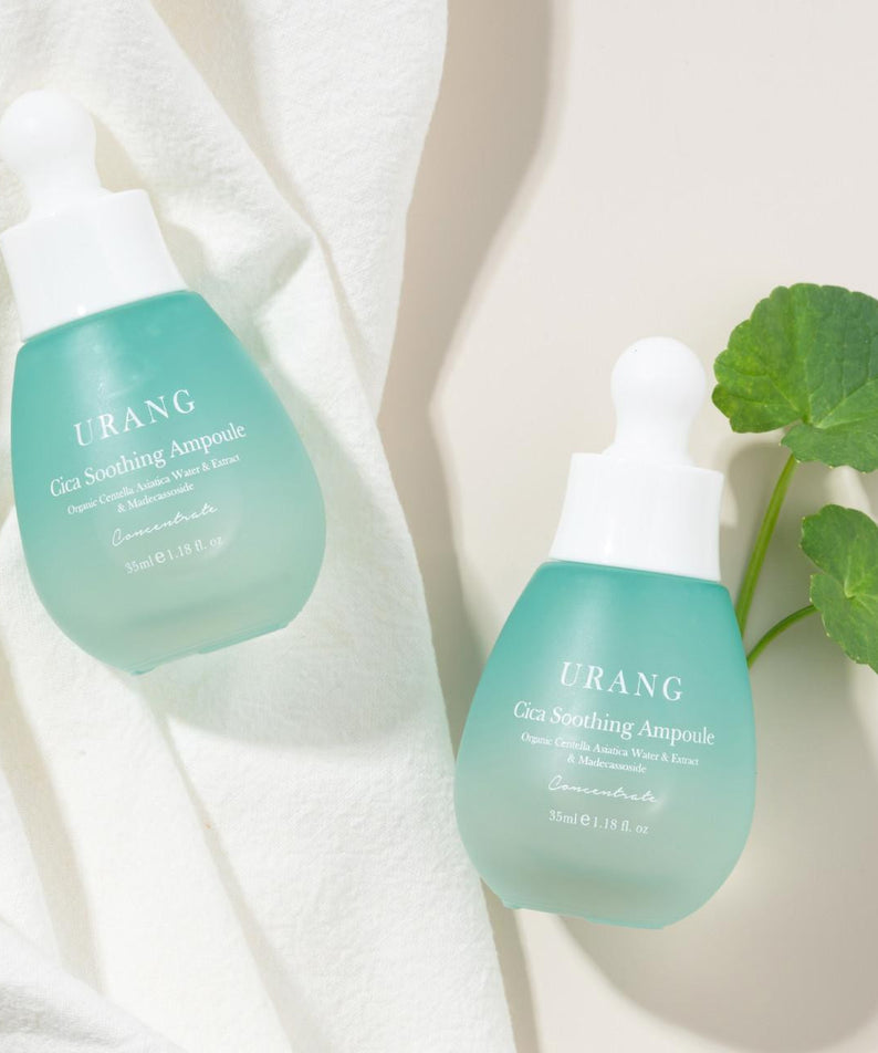 Deux flacons de URANG Cica Soothing Ampoule, posés sur un tissu blanc avec une feuille verte.