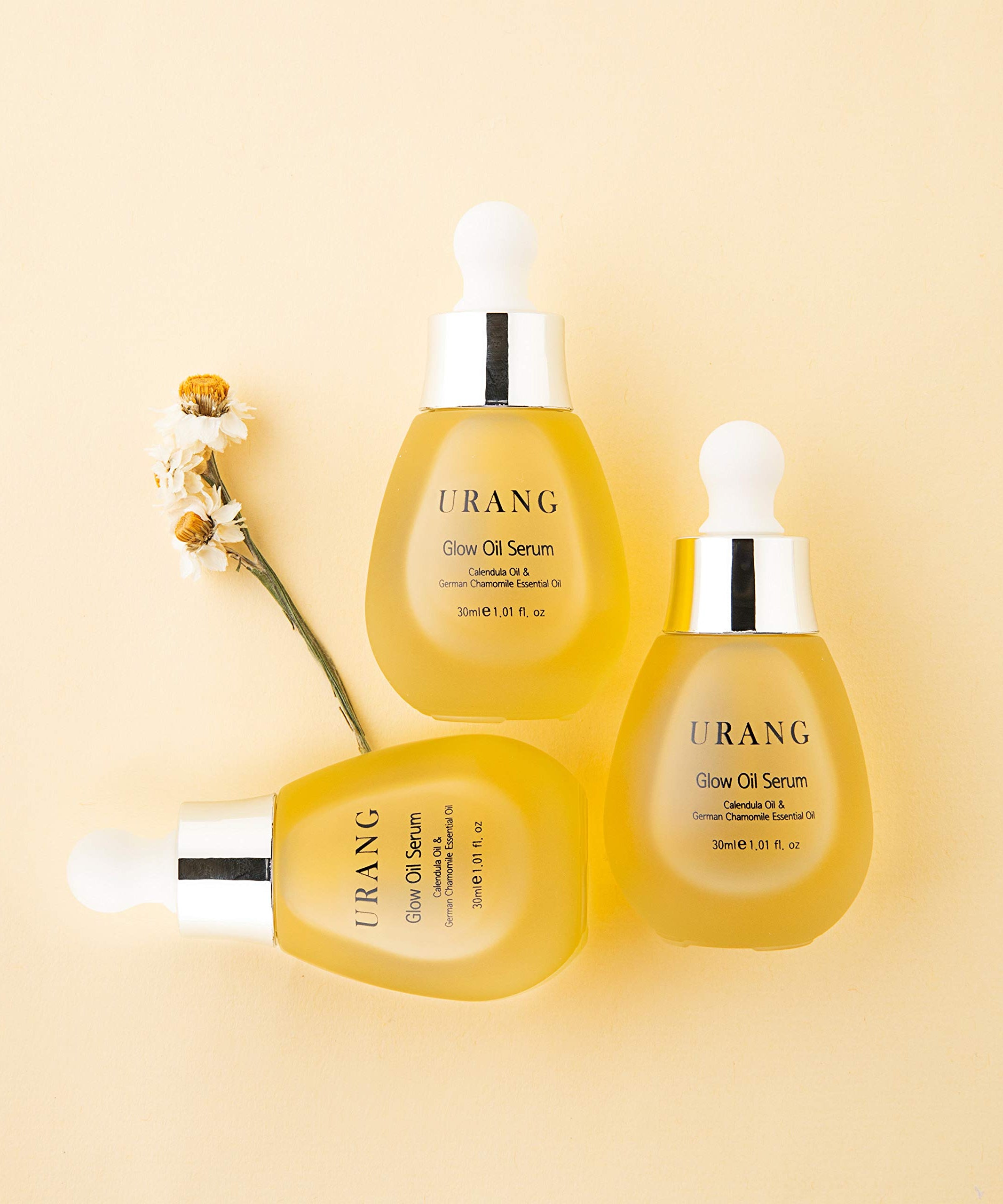Flacons de URANG Glow Oil Serum 30ml sur fond beige avec fleurs, soin visage éclatant.