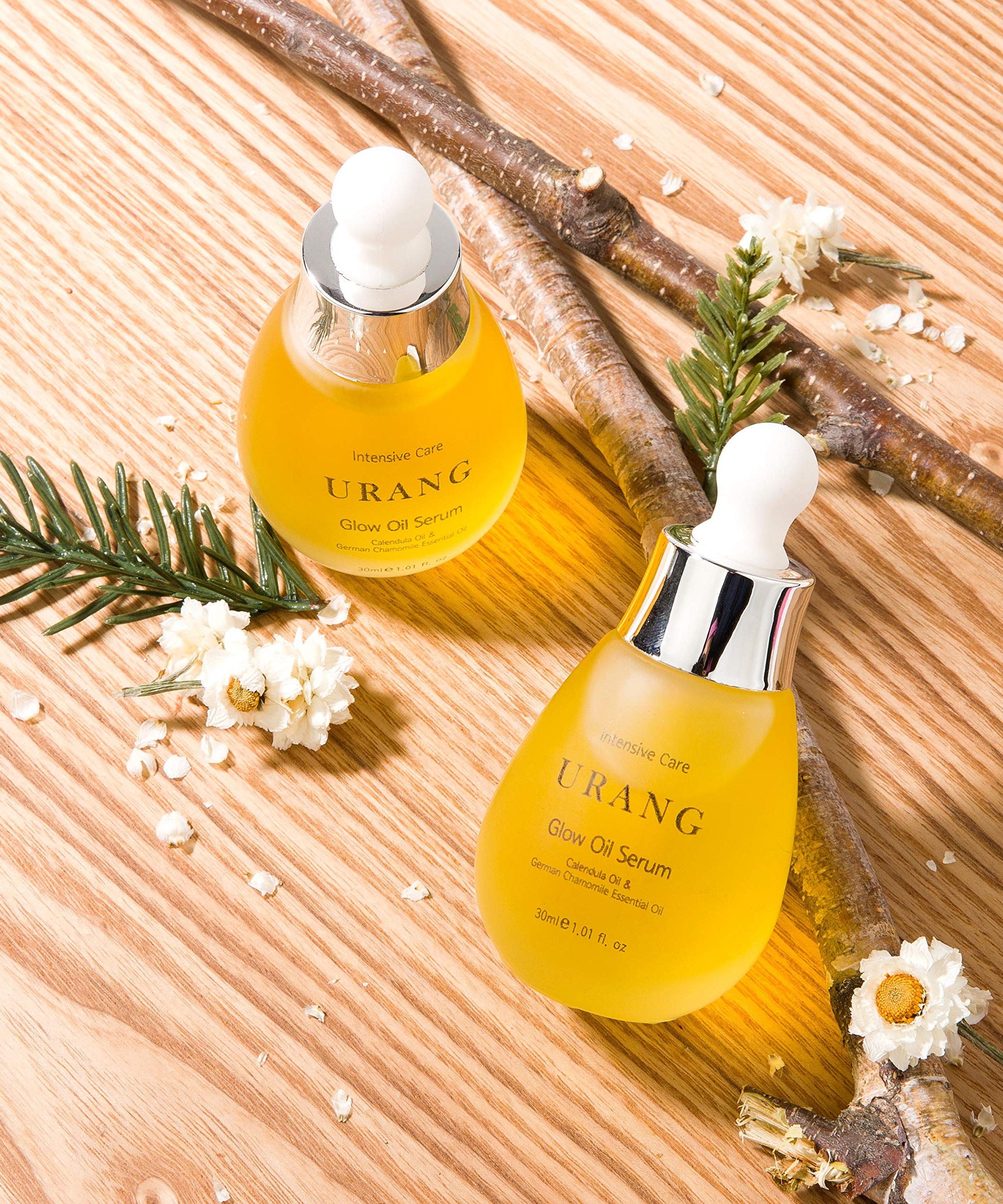 Bouteilles de URANG Glow Oil Serum 30ml sur fond en bois, entourées de branches de pin et de fleurs blanches.